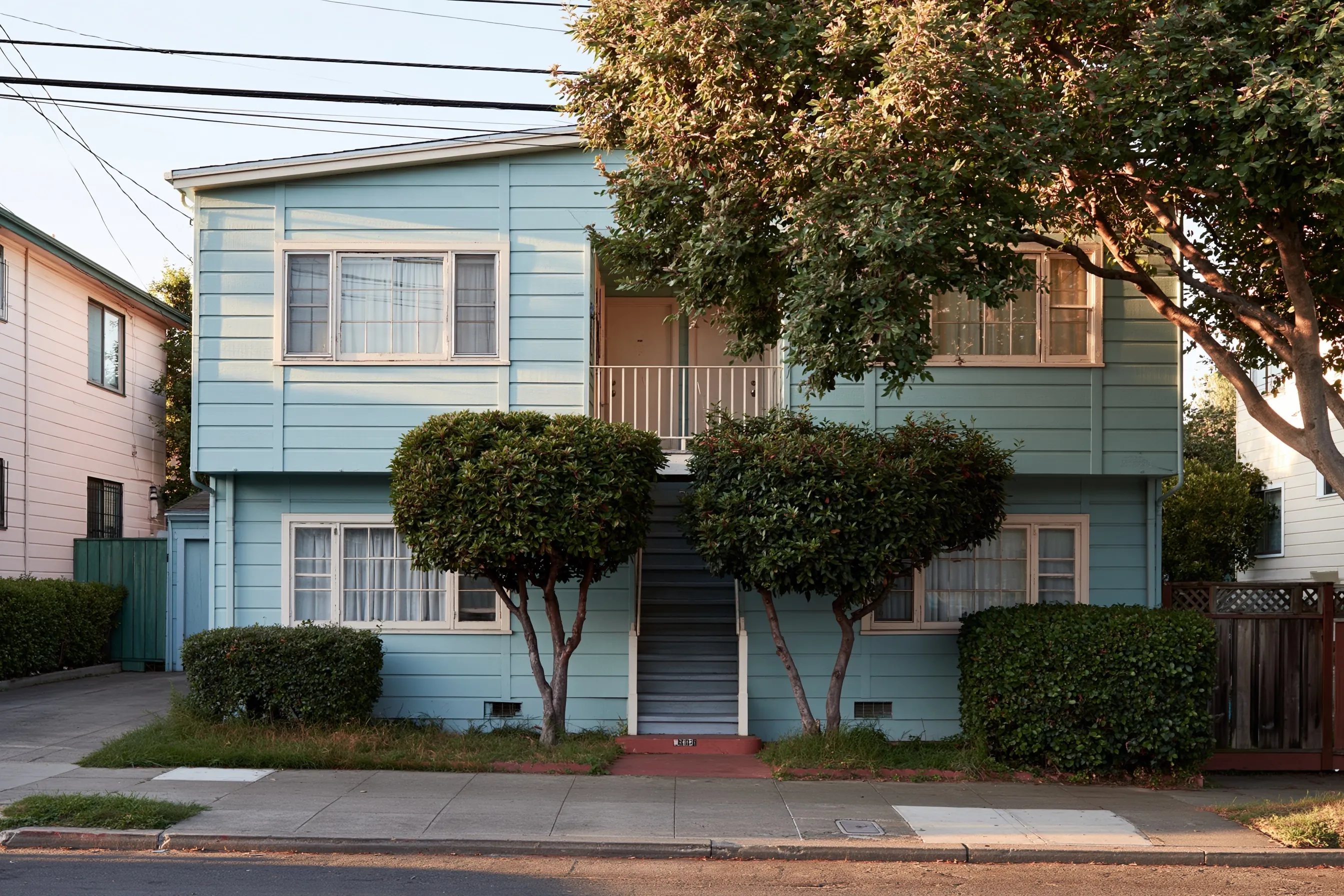 Two-story apartment building with painted wood siding, wood-framed windows and an exterior stair, two trimmed street trees in front